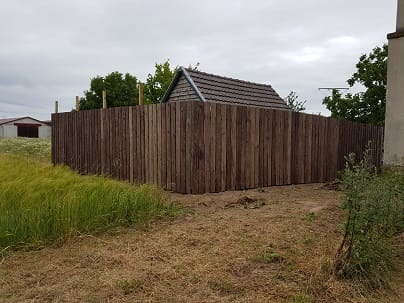 Der neue, hohe Holzbretterzaun steht am Rand der Wiese vor dem Häuschen mit Holzgiebel.